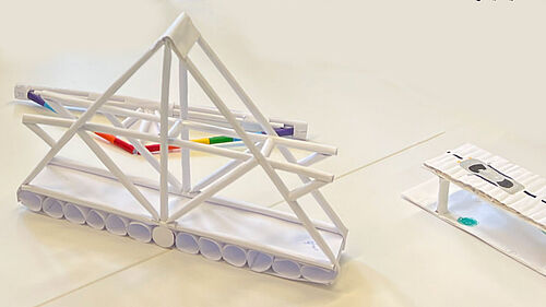 Es ist eine Papierbrücke aus weißem Papier zu sehen. Am rechten Rand befindetsich das Ende eier Paierbrücke auf die ein Auto gemalt wurde.