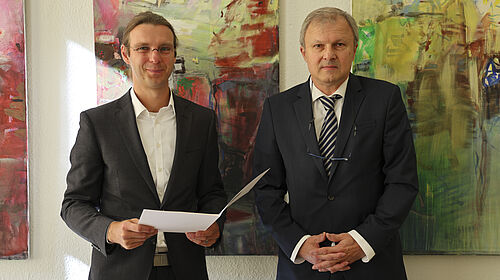 Ernennung Der Professor hält eine Mappe in der Hand und schaut in die Kamera. Der Gratulant steht neben ihm.
