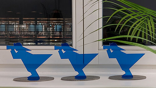 Die drei blau gefärbten Pokale in Form des Fischer-Signets aus dem Hochschul-Logo stehen auf einem Fensterbrett. Durch das Fesnter hinweg kann man den dunklen Himmel und die erleuchtete Bibliothek sehen.