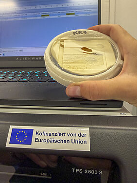 Eine Hand hält eine weiße runde Schale mit einer kleinen braunen Probe vor einem Laptop mit beleuchteter Tastatur, darunter ein Aufkleber mit dem Text 'Kofinanziert von der Europäischen Union'