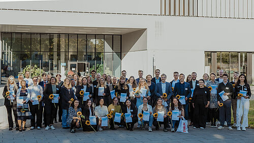50 Personen stehen neben- und hintereinander. Einige halten Blumen und Urkunden in den Händen. Daran erkennt man die Studierenden. Im Hintergrund ist Haus 6 zu erkennen.