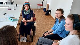 Eine junge Frau mit Halstuch und buntem Haarband sitzt im Rollstuhl und drei weitere junge Frauen sitzen dabei. Eine Frau trägt Kopfhörer.