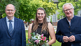  Drei Personen stehen auf dem Wismarer Campus, auf dem im Hintergrund Bäume mit frischem Grün zu sehen sind. Die mittlere Person ist die gewählte Prorektorin. Sie hält einen Blumenstrauß, alle drei tragen formelle Kleidung.