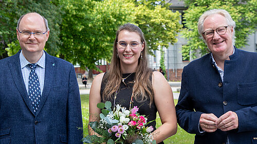 Drei Personen stehen auf dem Wismarer Campus, auf dem im Hintergrund Bäume mit frischem Grün zu sehen sind. Die mittlere Person ist die gewählte Prorektorin. Sie hält einen Blumenstrauß, alle drei tragen formelle Kleidung.