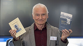 Schott Der Professor schaut in die Kamera und hält in jeder Hand ein Buch hoch.
