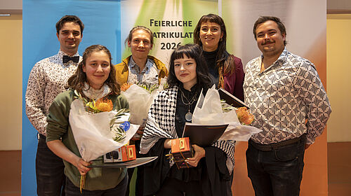 Sechs Personen stehen vor zwei Bannern mit Text, zwei Frauen halten Blumensträuße und Urkunden, alle lächeln in die Kamera.