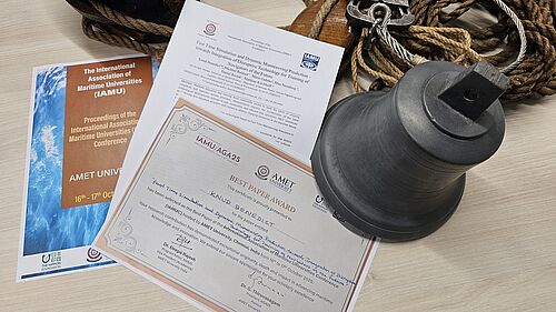 Die Auszeichnungsurkunde liegt auf dem Titelblatt des Konferenzbeitrags und dem Titelblatt des Konferenzbandes. Dahinter befinden sich Taue und eine Schiffsglocke.