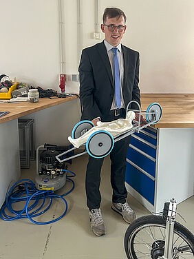 Pascal Schütt steht vor einer Werkbank. M it beiden Händen hält er ein rund 40 Zentimeter breites Modell in der Hand. In diesem Modell liegr eine ebenfalls im 3D-Drucke ersellte Figur.
