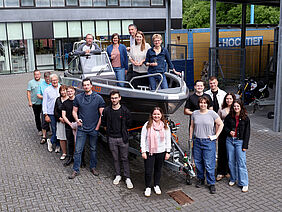 Personen stehen auf und um ein kleines Boot herum, welches noch "trocken" auf einem Trailer liegt.