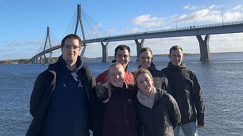Die Gruppe der sechs Studierenden steht am Ufer der Ostsee nähe der Stadt Vaasa. Im Hintergrund ragt die Raippalouto-Brücke, mit ihren 1045 Metern Gesamtlänge die längste Brücke Finnlands, empor.
