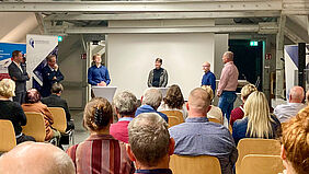 Über die Köpfe des Publikums hinweg sind sechs Personen zu sehen, die im Halbkreis stehen. Eine PErson sprciht, die anderen schauen sie an.