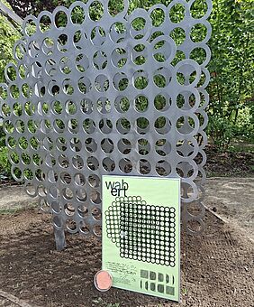 Das Bild zeigt das Kunstwerk WABBERT, welches eine Installation aus zwei aufrecht stehenden Metallplatten ist, die mit einer gleichmäßig verteilten Lochstruktur versehen sind.