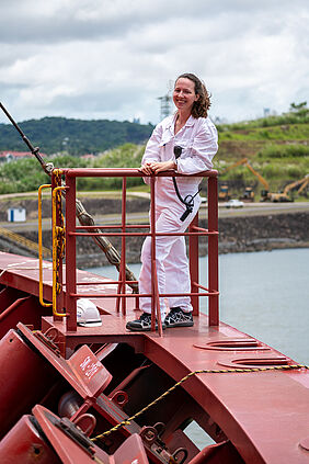 Verena lächelt in die Kamera, während sie in einen weißen Overall gekleidet an Bord eines Schiffes, das rot gestrichen ist, steht. Im nahen Hintergrund ist Land zu erkennen.