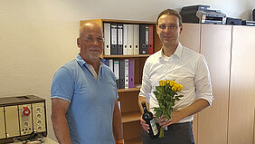 Der Professor hält einen Blumenstrauß und eine Weinflasche in der Hand Neben ihm steht Prof. Simanski. Im Büroschrank hinter den beiden befinden sich nur wenige Ordner.